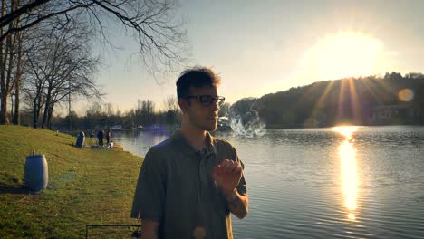 Hombre-De-Pie-Junto-A-Un-Lago-Fumando-Mientras-El-Sol-Se-Pone-En-La-Distancia