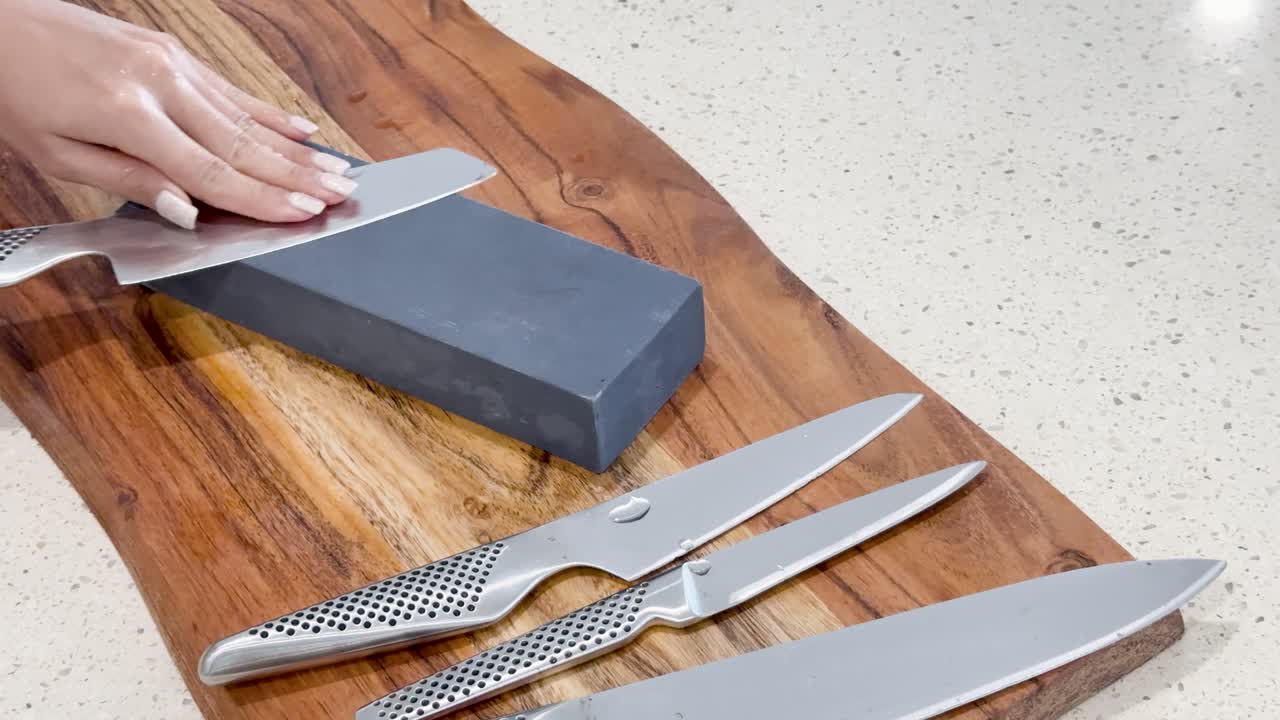 Hands methodically sharpen stainless steel kitchen knives on a whetstone atop a wooden board, under bright, even lighting with steady overhead camera angle
