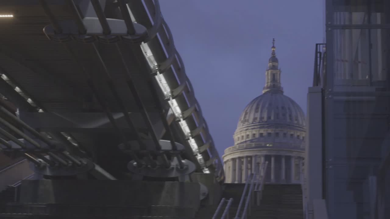 Evening city view of St Paul's Cathedral framed under the steel structure of Millennium Bridge as city lights glow along the riverside