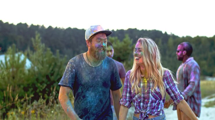 Young Cheerful And Smiled Multiethnic Friends In Colorful Paints Walking Outdoor And Talking Joyfully At The Holi Festival