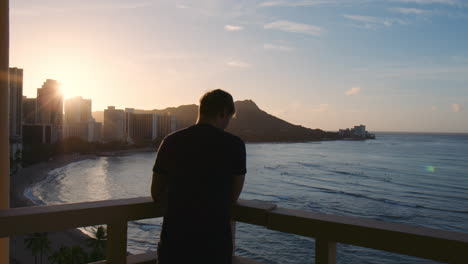 silhouetted tourist standing alone on hotel balcony watching beautiful morning sunrise over waikiki bay, hawaii