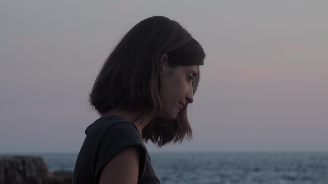 Ragazza seduta su una roccia in riva al mare con il tramonto sullo sfondo, rallentatore, primo piano