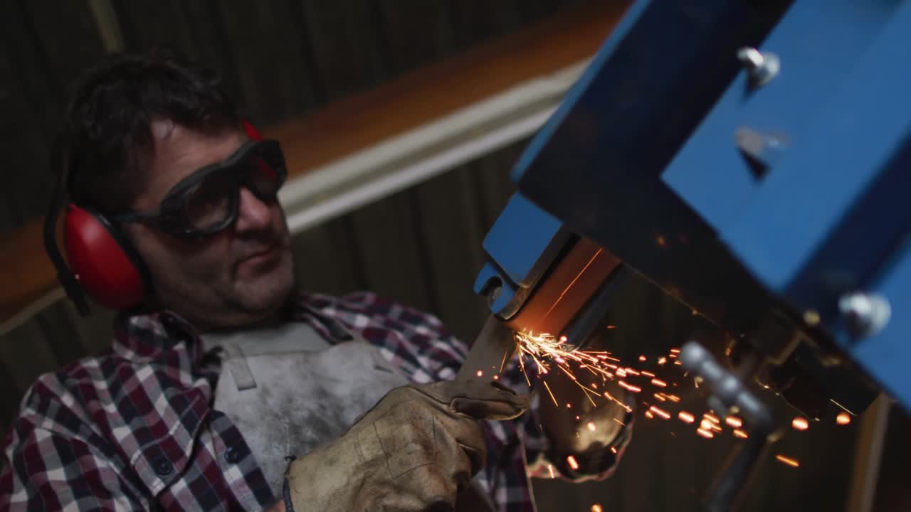 hombre caucásico fabricante de cuchillos en un taller con gafas y auriculares, usando un molinillo angular