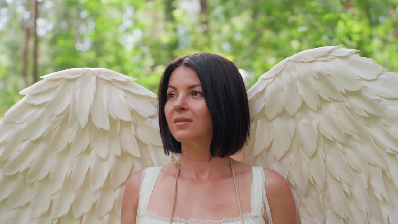 Lady dressed in angelic costume with large white wings standing in lush green forest, calm expression and soft gaze reflecting peace, grace, and divine presence under sunlight