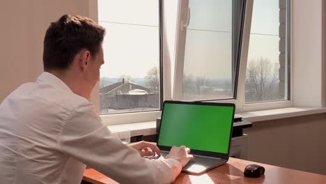 a young man enjoys success at work sitting at a desk in front of a computer with a green screen. victory, success in work