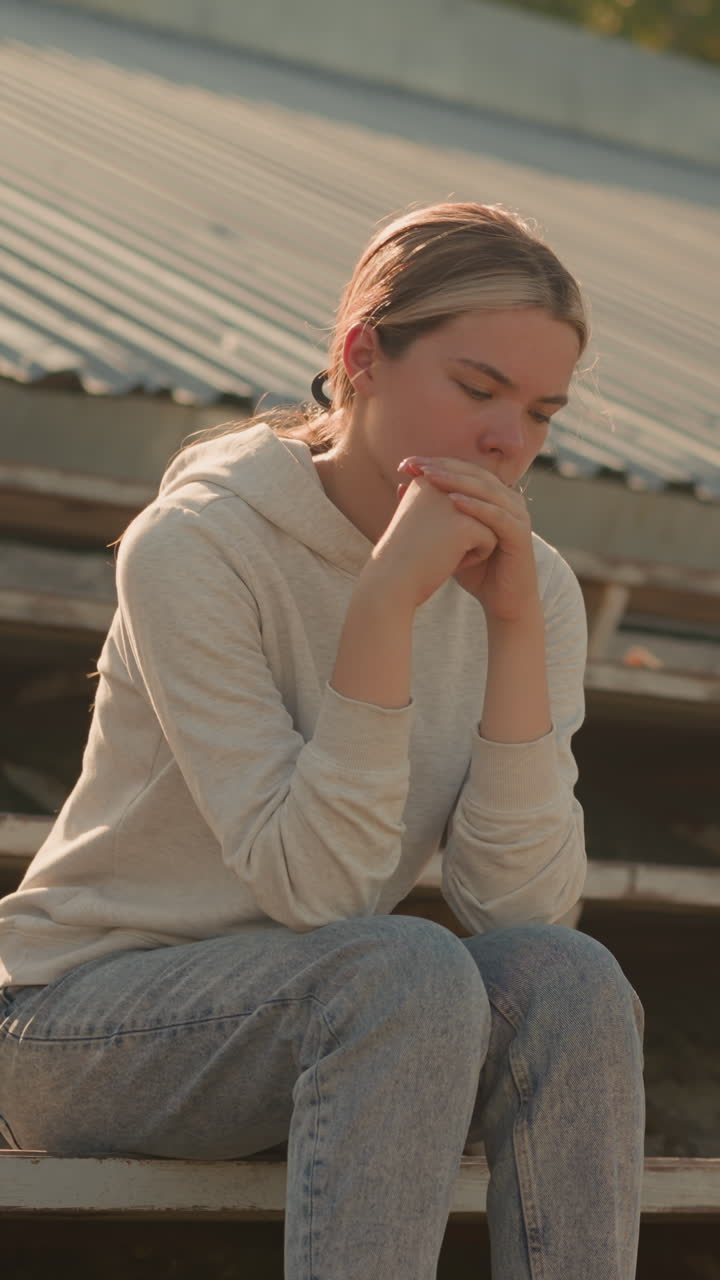 Woman in casual hoodie and jeans sits thoughtfully on rustic stadium bleachers, hands joined together, with a reflective expression