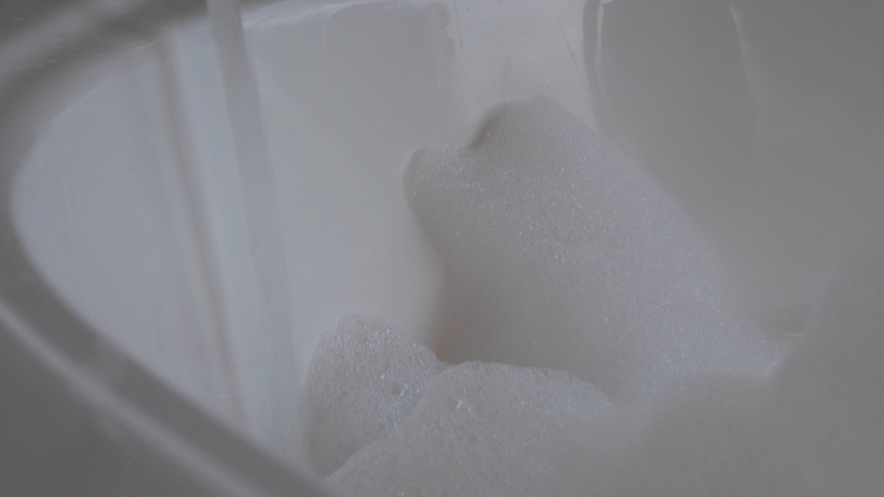 Slow motion pan down of tap running hot water into white bubble bath with foam build up, beautiful home getaway with luxury relaxation.