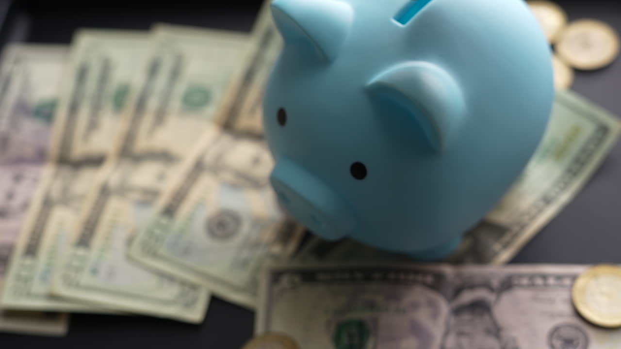 Primer plano detallado de una hucha azul rodeada de billetes de dólar y monedas, que simboliza el ahorro y la creación de un fondo de emergencia. La iluminación suave destaca el tema financiero.
