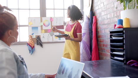 Diverse-female-fashion-designers-using-laptop-and-tablet-discussing-designs-at-studio,-slow-motion