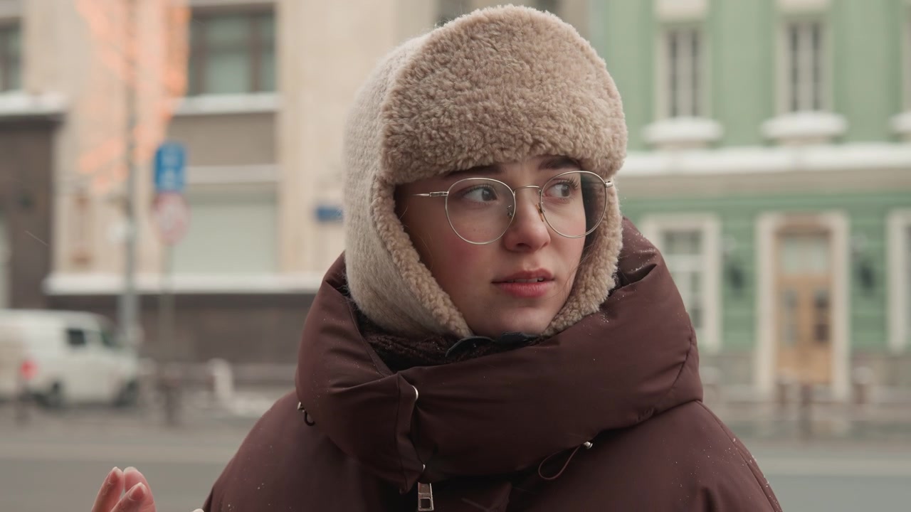 Ritratto di donna di tre quarti posteriore che indossa un cappello da trapper in maglia e un cappotto invernale imbottito, che gira la testa e guarda a destra mentre la neve cade e un veicolo sfocato si muove nel traffico urbano in una fredda luce nuvolosa