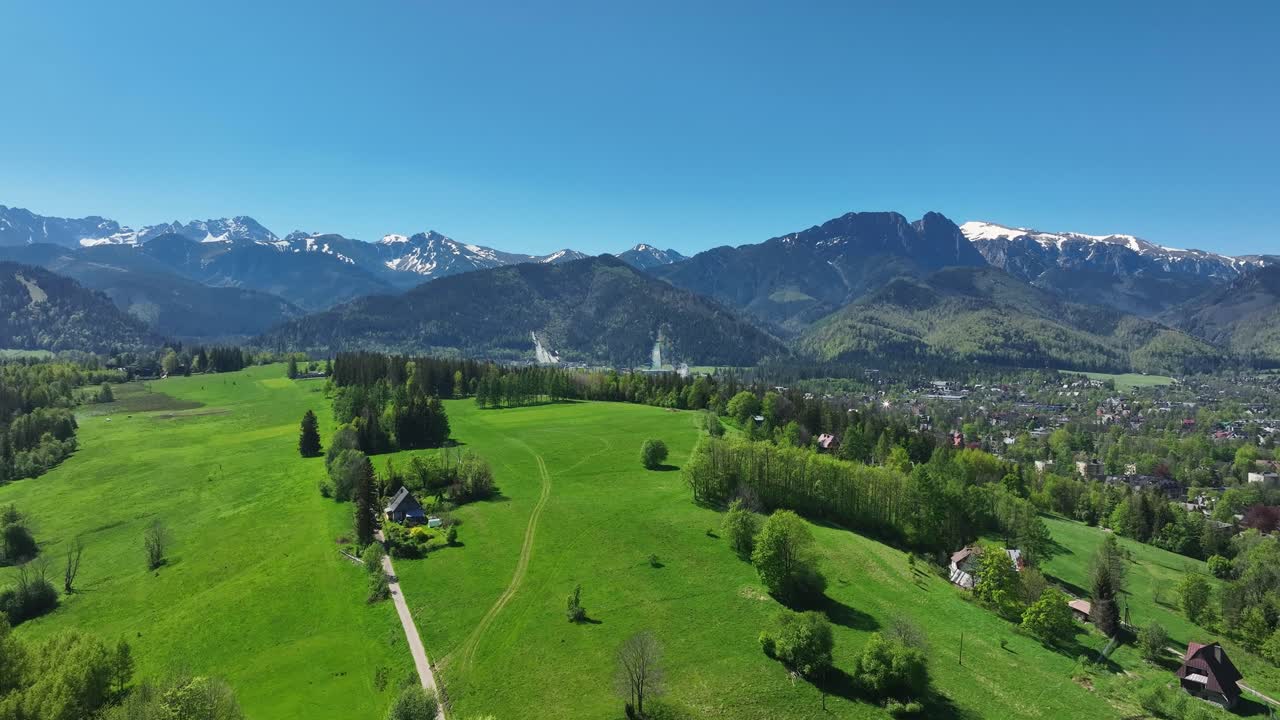 Aerial View of a Lush Green Valley with Snow-Capped Mountains and a Village Under a Clear Blue Sky