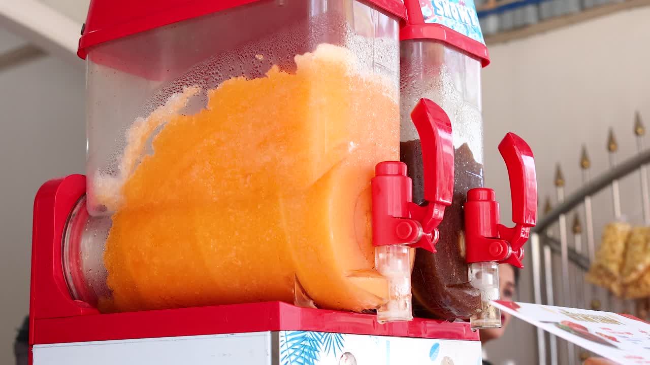 A vibrant orange slushy machine operates in a bustling Phuket market, with bright lighting and dynamic camera angles capturing the scene