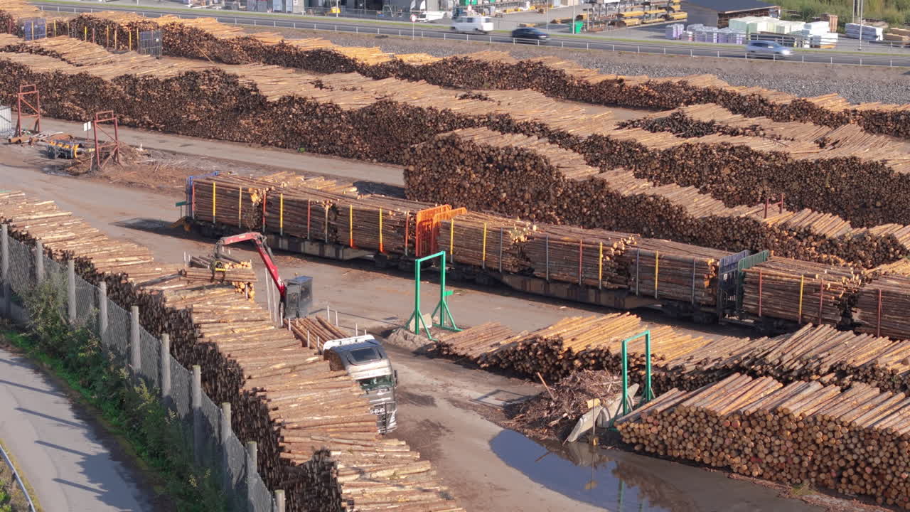 Side aerial view of grapple log loader unloading timber logs from truck at yard
