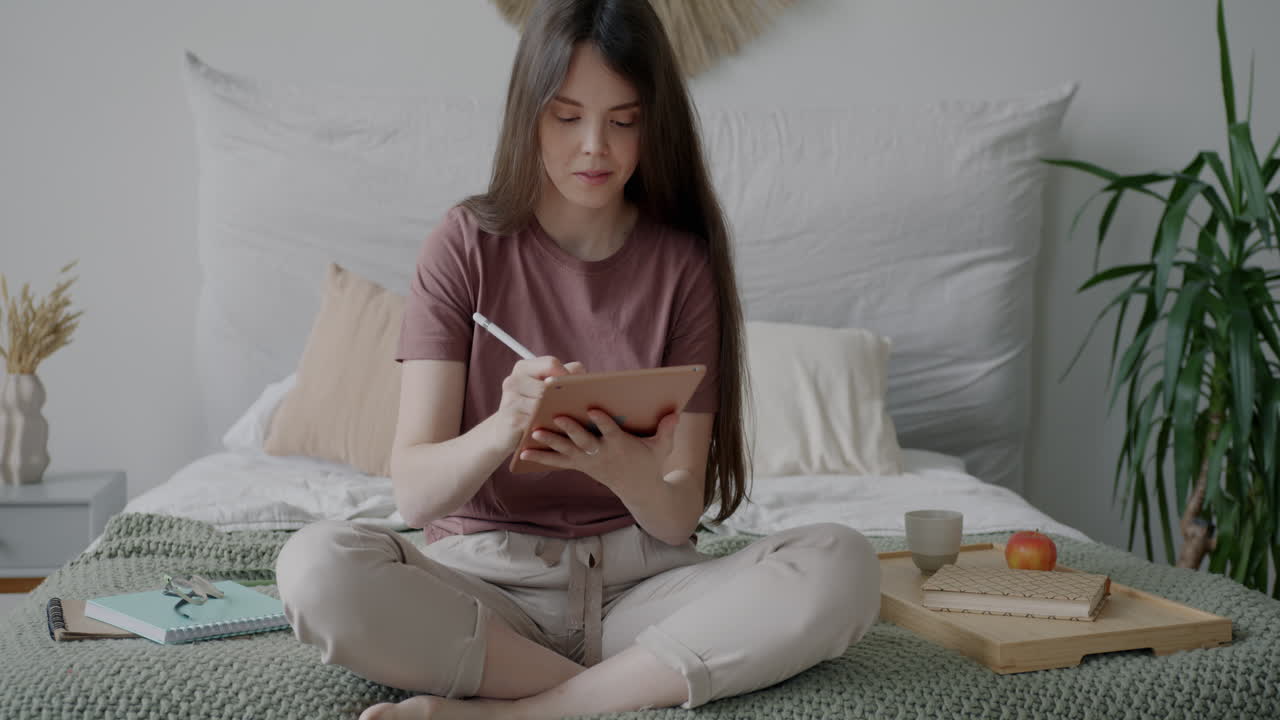 Woman working on digital tablet in a cozy bedroom.