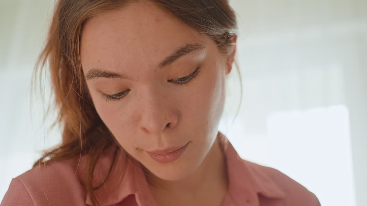 Young Caucasian Woman Looking Down In Soft Morning Light, Contemplative Expression While Reading Journal By Window, CloseUp Portrait, Natural Makeup, Pink Shirt, Intimate Home Interior, Slow Living