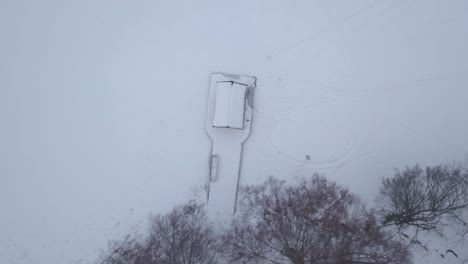 Overhead-panning-from-right-hand-side-revealing-tiny-shed-on-edge-of-frozen-lake-close-to-deciduous-tree-line