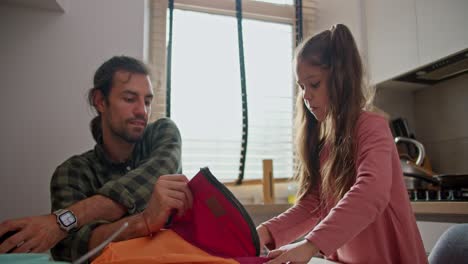 una pequeña chica morena en un vestido rosa, junto con su padre y un hombre moreno en una camisa a cuadros verdes, recoge su brillante mochila de color naranja-rojo para la escuela para la primera lección, en la cocina de un apartamento moderno