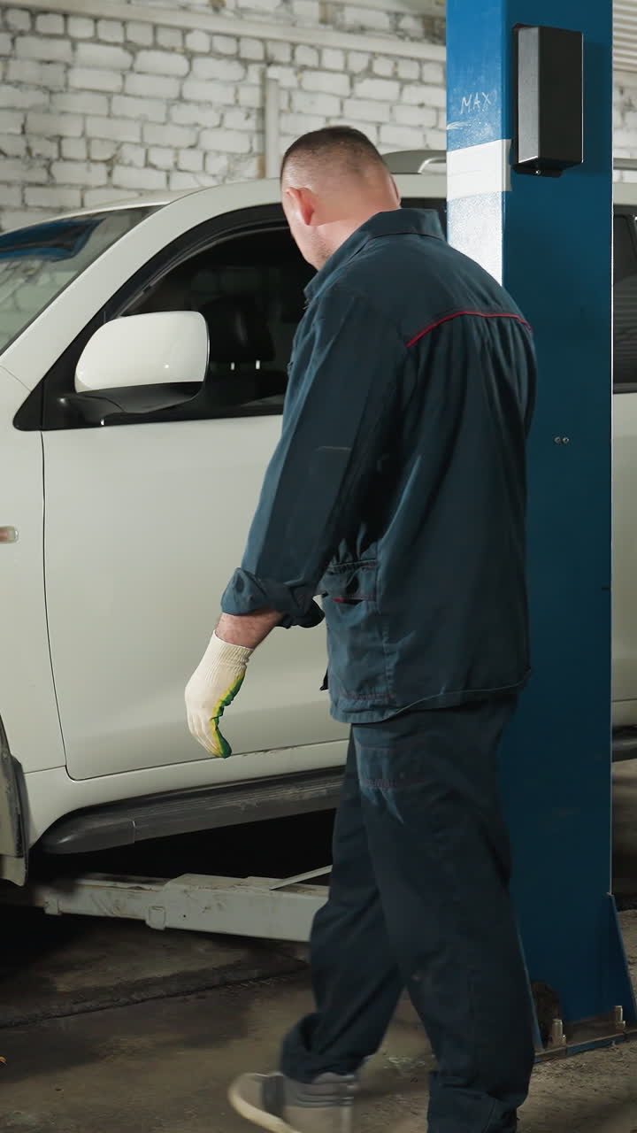 파란 유니폼을 입은 기계공이 작업장에서 자동차를 들어 올리는 것을 검사하고 관찰하며 차량 세부 사항에 초점을 맞추고, 배경에는 장비와 도구가 보입니다.