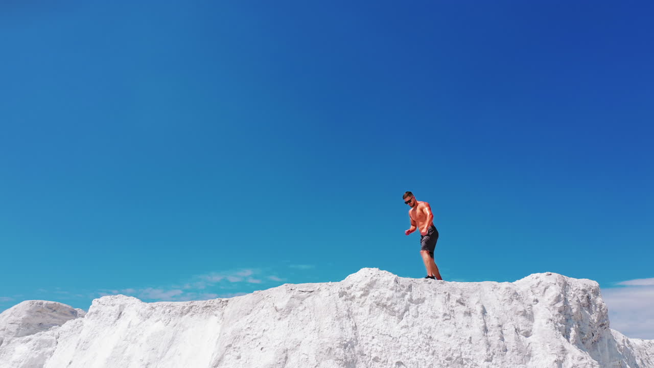 Handsome man in shorts with muscular body standing on the white hill under blue sky. Bodybuilder without shirt posing on the top of the mountain. Drone view.