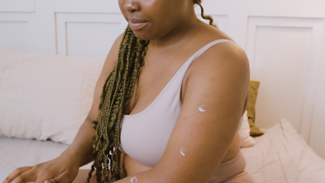 Close-up view of African american woman in underwear sitting on the bed spreading moisturizing cream on her arms