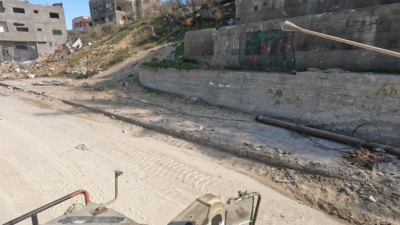 Punto di vista sulla Striscia di Gaza da un veicolo militare che si muove attraverso un'area urbana devastata con edifici distrutti e un grande muro di cemento coperto di graffiti arabi