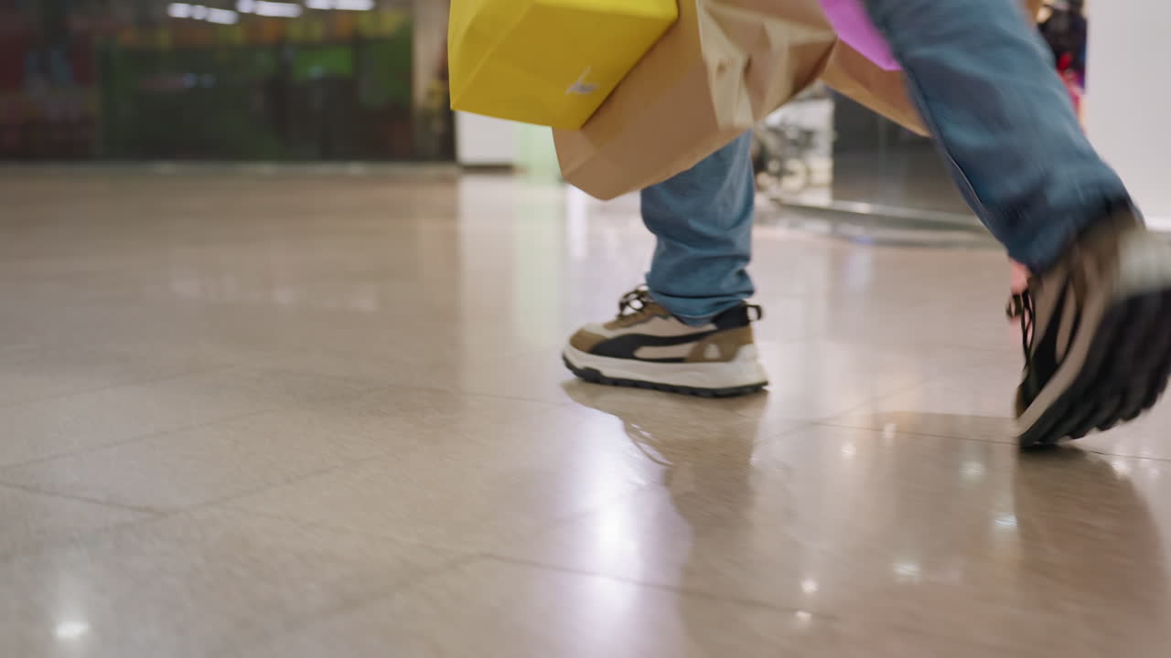 Leg view of man in jeans and sneakers walking on shiny floor carrying paper shopping bags inside brightly lit shopping center with modern retail environment