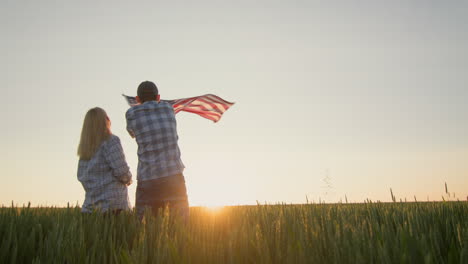 an energetic couple waving the american flag in front of a field of wheat as the sun sets. 4th of july concept