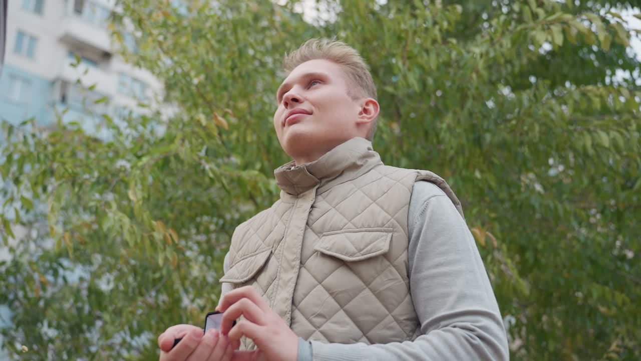White man kneels with warm smile holding ring box as he proposes to girlfriend outdoors with green leaves swaying gently in wind and blurred urban buildings in background