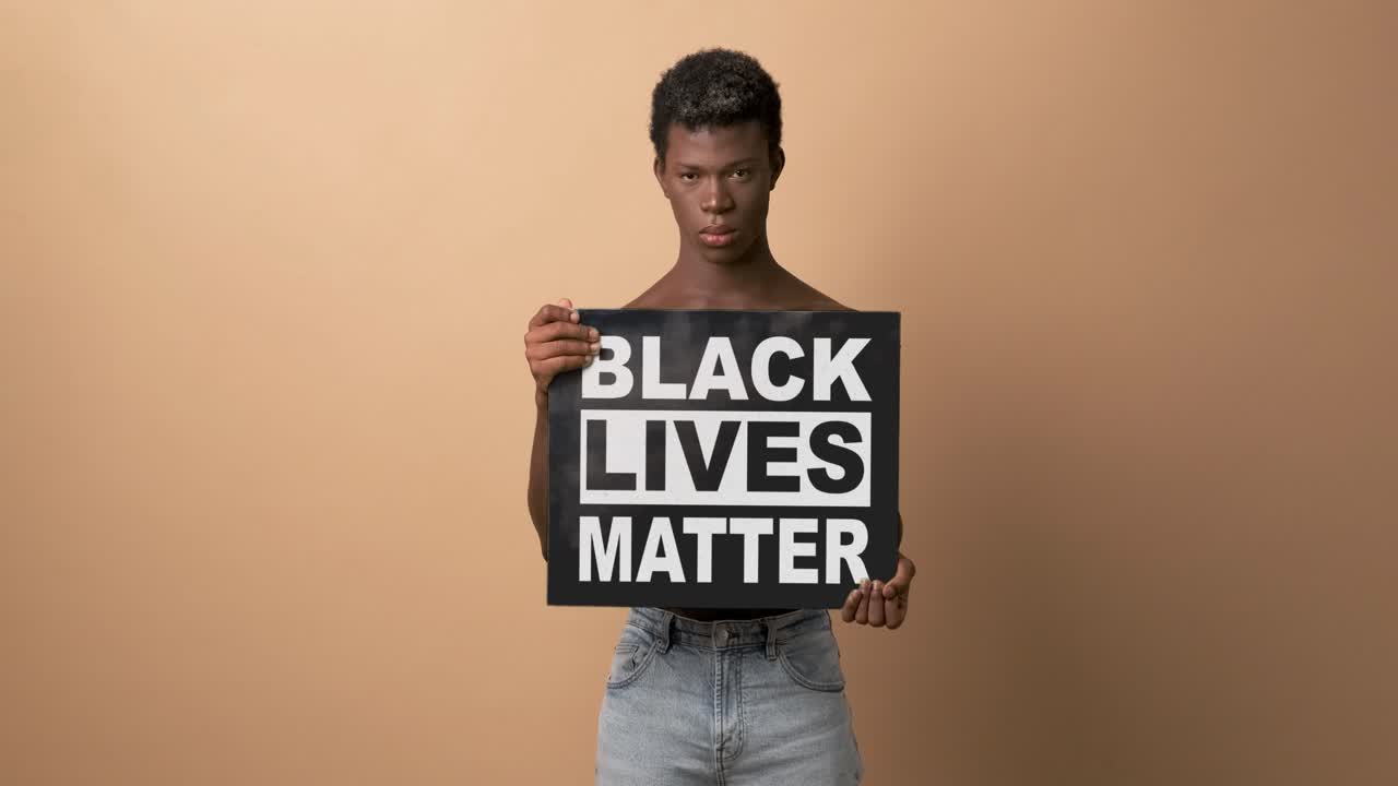 Black shirtless man holding a sign with the phrase BLACK LIVES MATTER looking angrily at camera