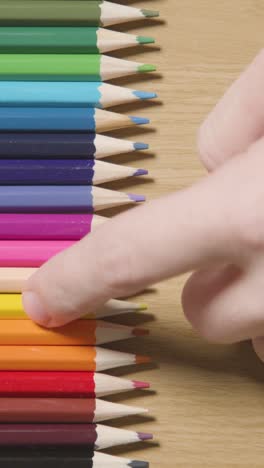 video vertical de lápices de colores dispuestos en una línea sobre un fondo de madera con una persona que elige uno amarillo