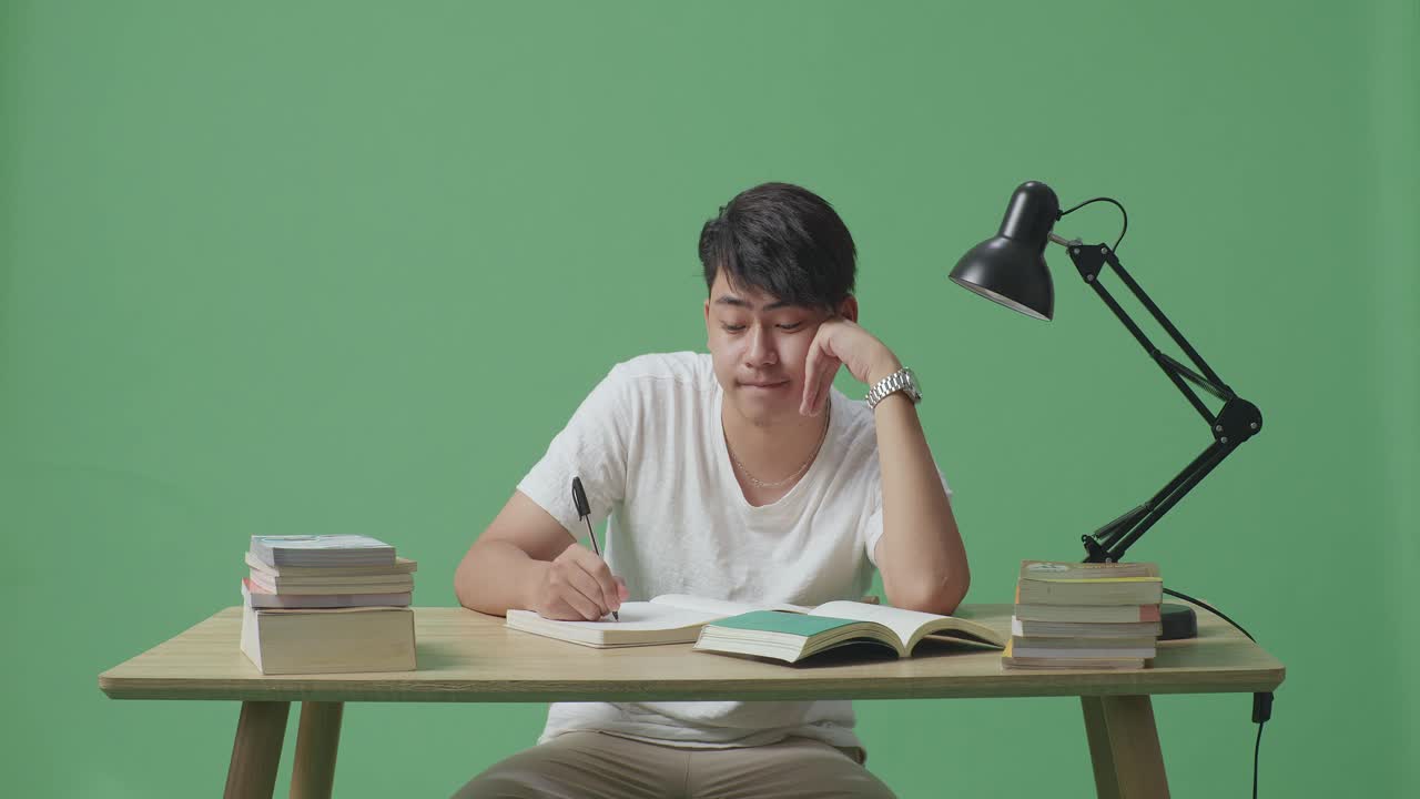 젊은 아시아 남성이 집에서 녹색 화면 배경에서 공부하는 동안 책에 쓰는 것에 지루합니다.