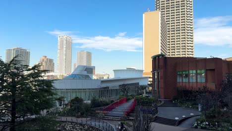 urban park with modern buildings backdrop, clear sky, lush greenery, and red bridge, daytime, calm city vibe