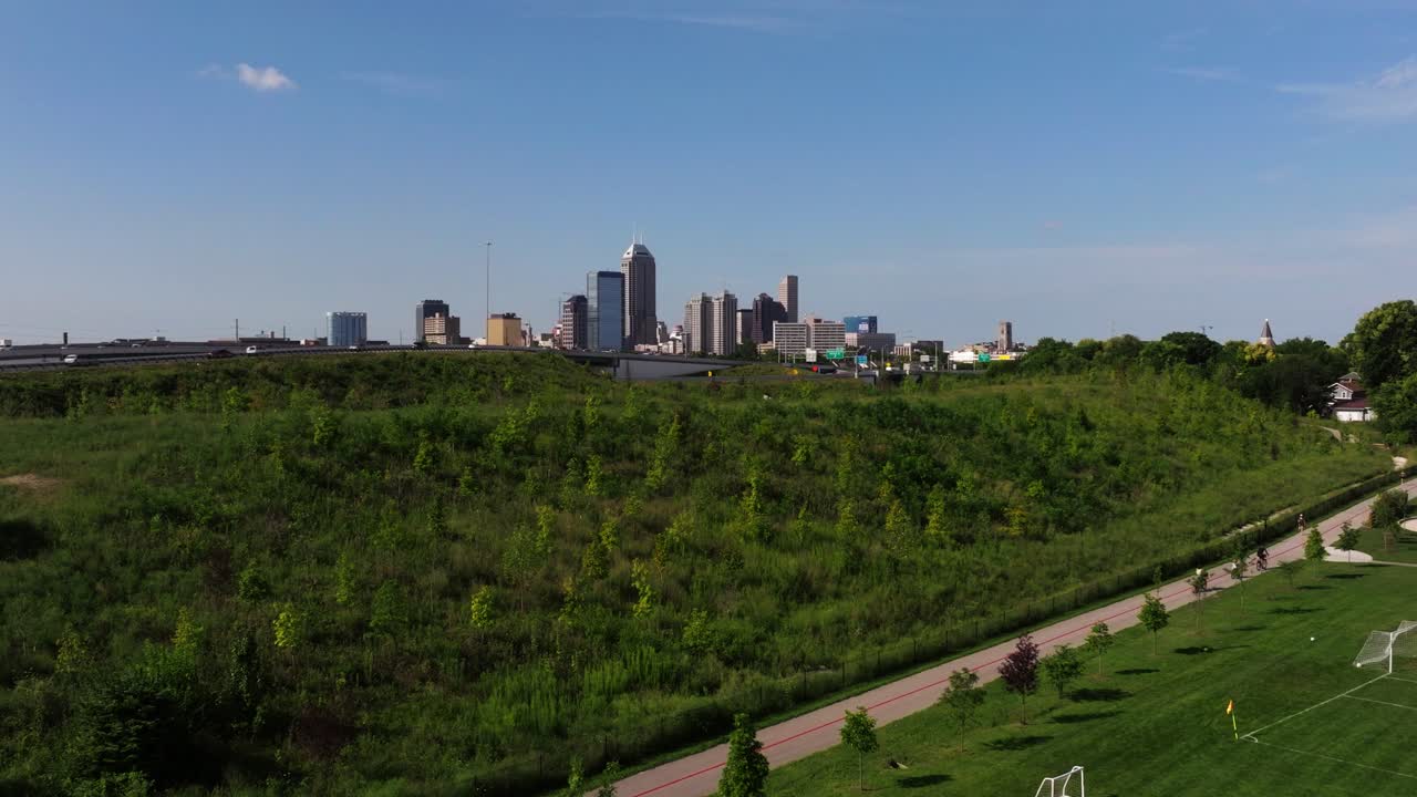 Drone holds a static position, framing a green hill and park with downtown Indianapolis skyline on the horizon, leaving generous copy space