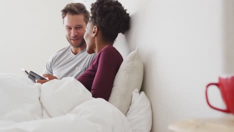 video of happy diverse couple using tablet in bed and talking