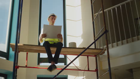 a young worker studies a drawing, sits tall on scaffolding