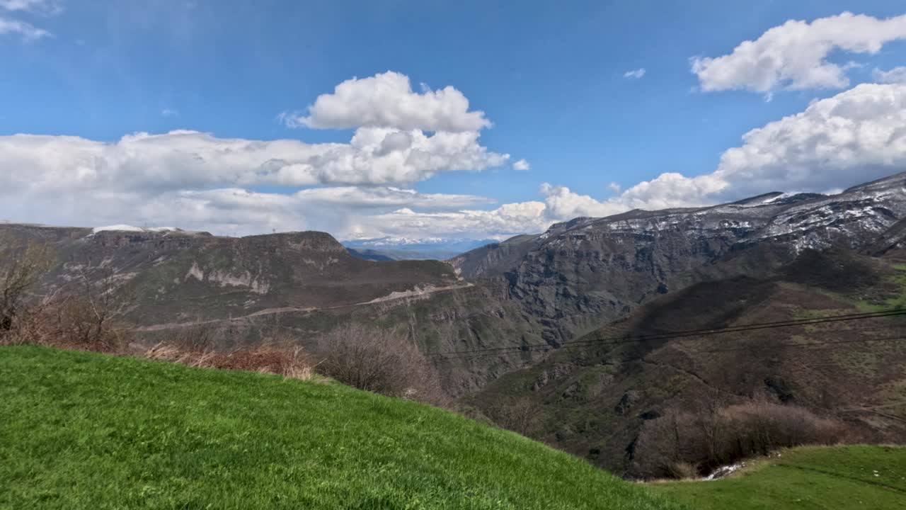 Le persone osservano un vasto paesaggio di montagne e valli. Erba verde copre il terreno mentre le nuvole fluttuano sopra. La neve è visibile su alcune cime in lontananza, segnando il cambio di stagione.