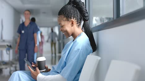 happy african american female doctor using smartphone, drinking coffee in corridor, slow motion