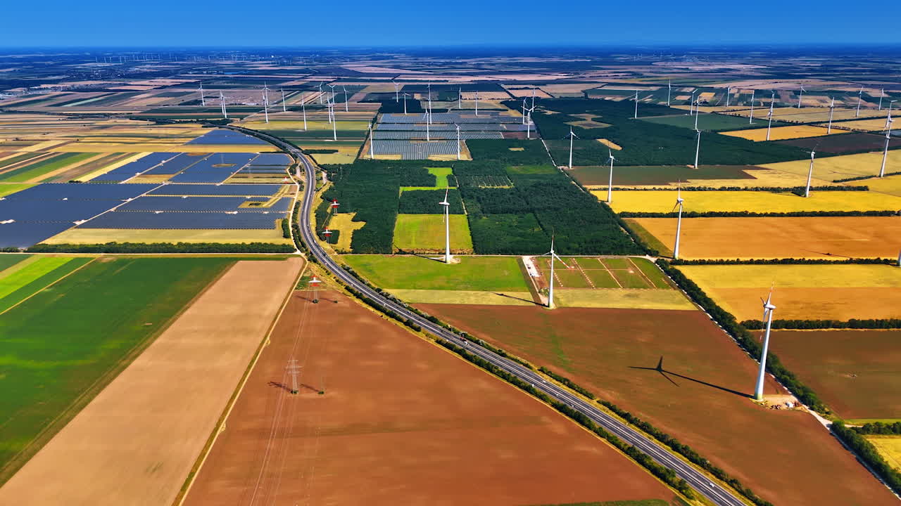 Highway crosses the vast agricultural plantations. Solar panels sites and wind turbines installed in the field. Aerial view