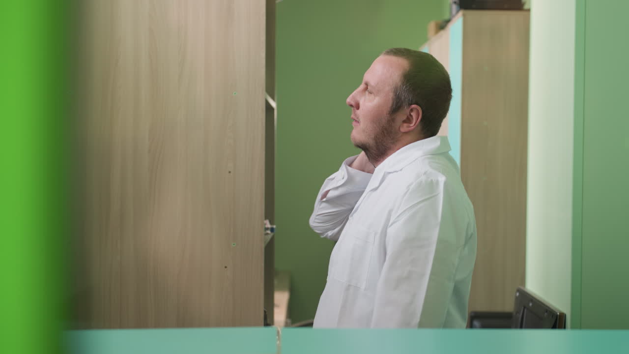 una vista lateral de un técnico de laboratorio en un abrigo blanco girando el cuello con la mano derecha y también estirando la mano, con dos estantes al lado de la caminata
