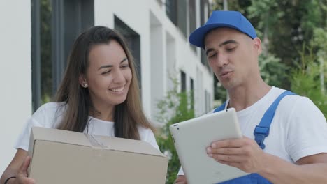 beautiful woman meets a delivery man who gives her a parcel box beside her home, she signs electronic signature on tablet. concept of courier, home delivery, online shopping