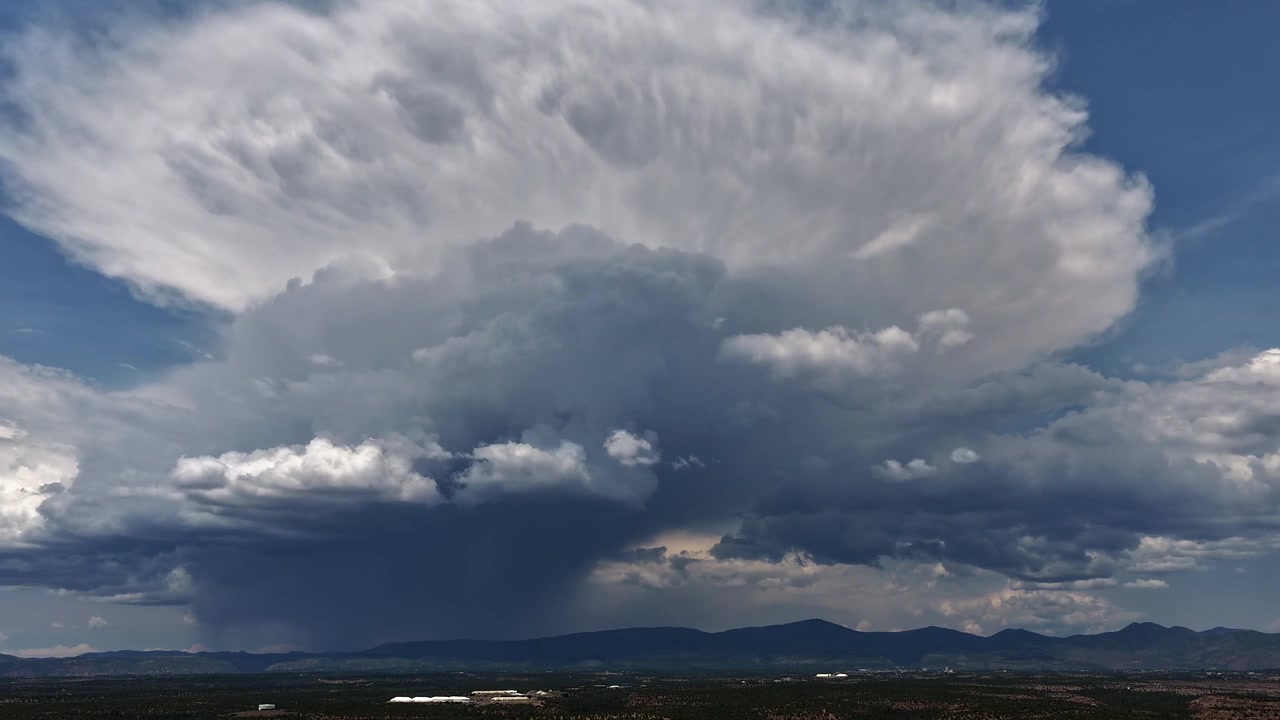 4K UHD drone-timelapse af en massiv monsunstorm, der skifter form over bjerge. En tårnhøj hvid sky med en mørk regnsøjle udvider sig dynamisk, hvilket illustrerer ekstremt klima og sommervejr