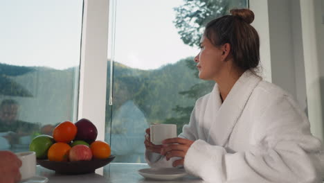 brooding wife in robe drinking tea near window. wife with hair tied back spends morning with cup of flavored coffee opposite husband. woman in bathrobe