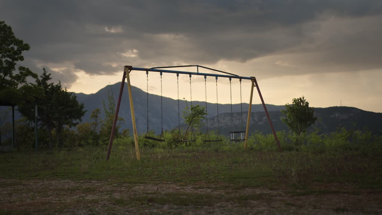 Static wide shot of an old, empty swingset in an overgrown clearing, with moody clouds and a mountain ridge silhouetted in the background.