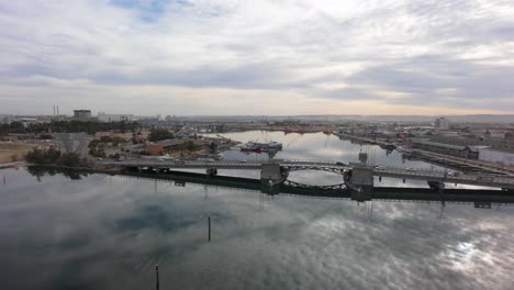 slow flight towards a draw bridge on port adelaide river with cars running over the bridge, clouds reflecting on the calm water with shipping facilities in the background
