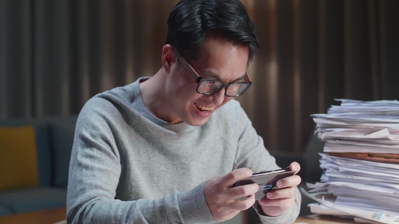 집에서 문서 작업을 한 후 스마트폰에서 우승 게임을 축하하는 행복한 아시아 남자의 클로즈업