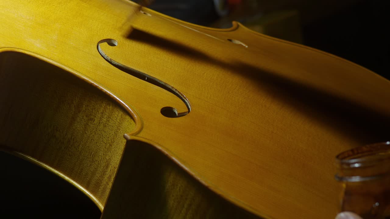 Artistic close-up captures a luthier’s hand applying varnish to a cello’s soundboard, accentuating the graceful f-hole and wood grain in the light of a Cremona workshop