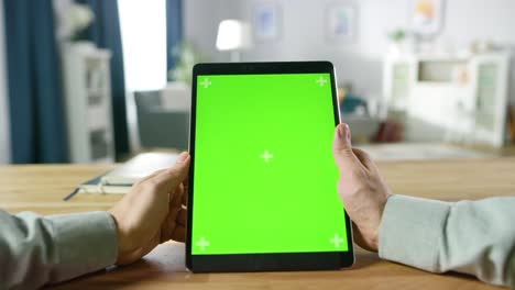 primer plano de un hombre usando gestos con las manos en una pantalla de maqueta verde, una tableta digital en modo retrato mientras está sentado en su escritorio. en el fondo, una acogedora sala de estar.