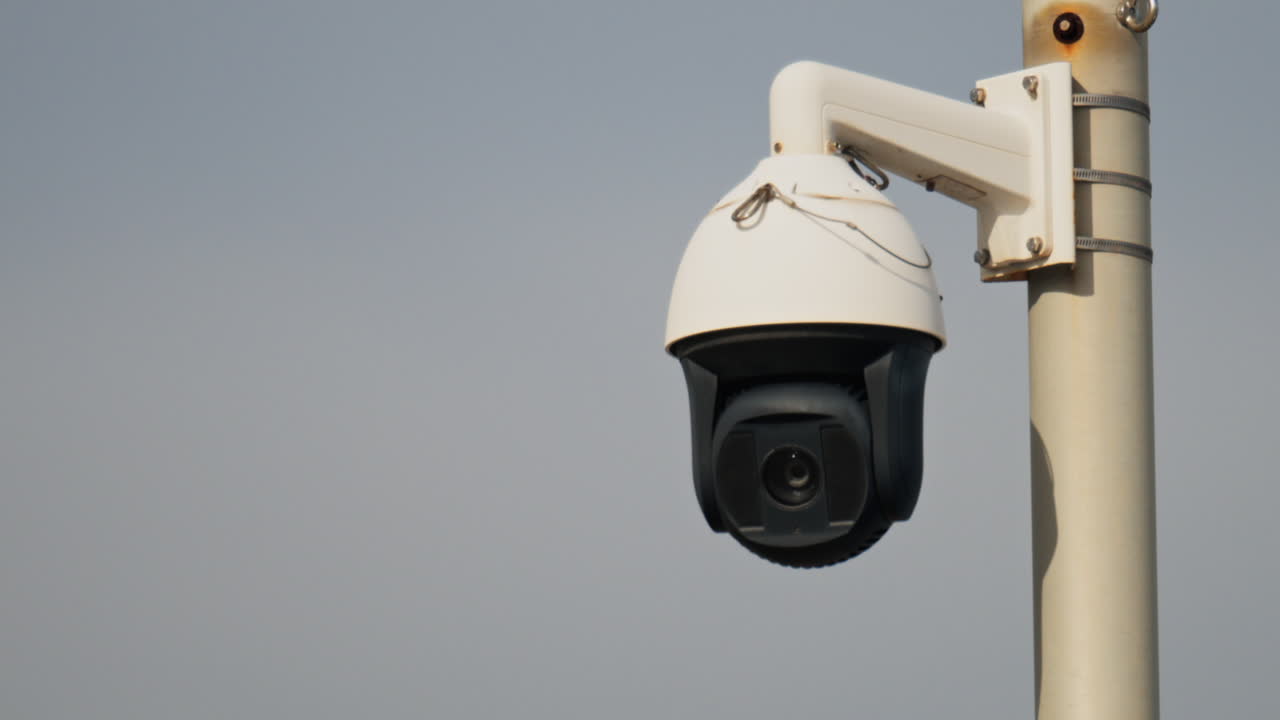 Close up of a round, black and white surveillance camera mounted on a white pole with the cloudy sky on the background