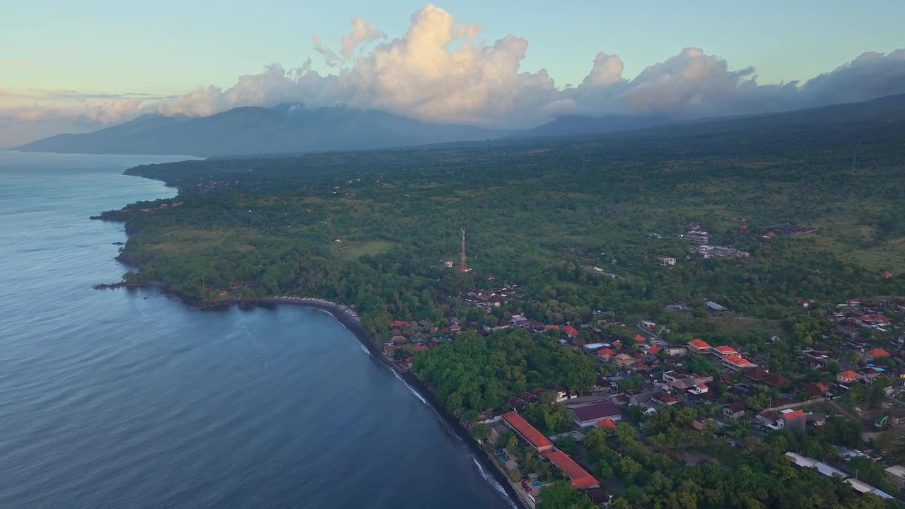 Rekaman drone udara garis pantai Tulamben di Bali menampilkan desa, laut, dan pegunungan saat senja.