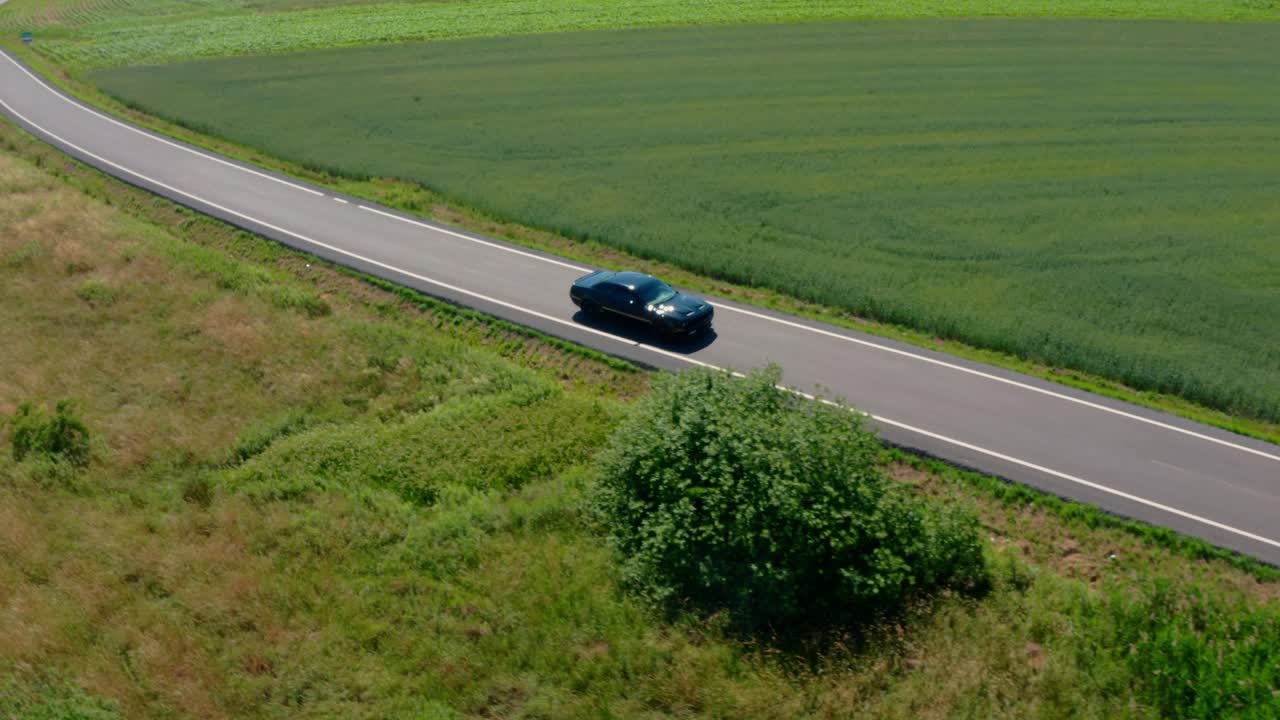 A drone shot showcasing a dark Dodge Challenger Scat Pack driving down an empty road through picturesque rural landscapes.